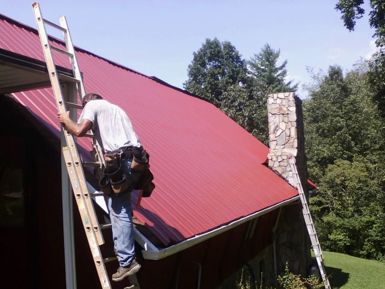 Roofer on a ladder beside a red metal roof and stone chimney — Soffit & Fascia Repair by Roofing Brothers in Boomer